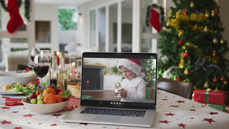 Happy-caucasian-boy-in-santa-hat-on-laptop-lying-on-christmas-table