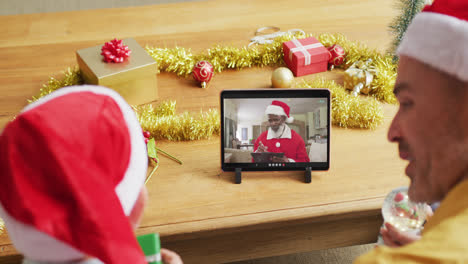 Caucasian-father-and-son-with-santa-hats-using-tablet-for-christmas-video-call-with-santa-on-screen