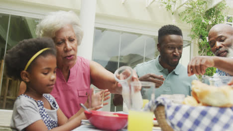 Happy-african-american-family-pouring-juice-and-having-breakfast-in-garden