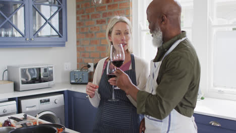 Mixed-race-senior-couple-wearing-aprons-toasting-wine-glasses-while-cooking-in-the-kitchen-at-home