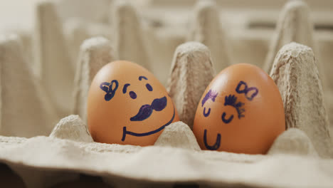 Video-of-close-up-of-two-brown-eggs-with-drawn-faces-in-egg-carton-background