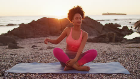 African-american-woman-practicing-yoga-and-meditating-on-the-rocks-near-the-sea-during-sunset