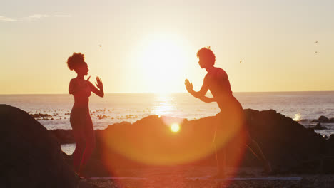 Afroamerikanisches-Paar,-Das-Bei-Sonnenuntergang-Gemeinsam-Yoga-Auf-Den-Felsen-In-Der-Nähe-Des-Meeres-Praktiziert