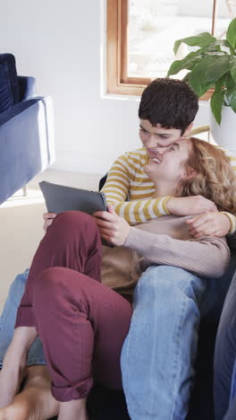 Happy-caucasian-lesbian-couple-lying-on-sofa,embracing-and-using-tablet-in-sunny-living-room