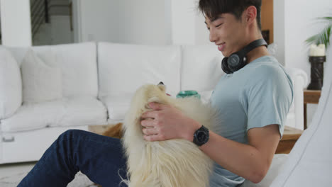 Glücklicher-Asiatischer-Männlicher-Teenager-Streichelt-Seinen-Hund-Und-Trägt-Kopfhörer-Im-Wohnzimmer