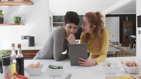 Happy-caucasian-lesbian-couple-standing-at-counter,-talking-and-using-tablet-in-sunny-kitchen