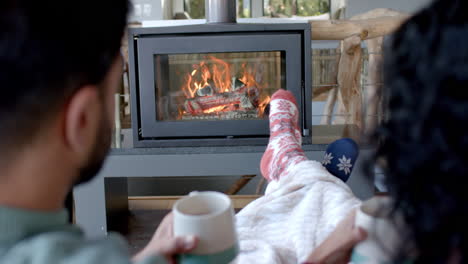 Happy-biracial-couple-on-sofa-in-warm-socks-under-blanket-in-front-of-fireplace-at-home,-slow-motion