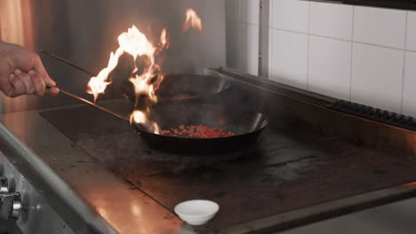 Caucasian-male-chef-frying-food-in-frying-pan-with-bursting-fire-in-kitchen,-slow-motion