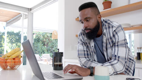 Hombre-Afroamericano-Tomando-Café-Y-Usando-Una-Computadora-Portátil-En-Una-Cocina-Soleada,-Cámara-Lenta