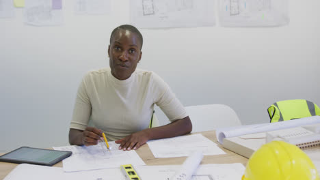 Portrait-of-happy-african-american-female-architect-sitting-at-desk-with-blueprints