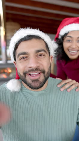 Vertical-video-of-biracial-couple-wearing-santa-claus-hats-having-video-call-at-home,-slow-motion