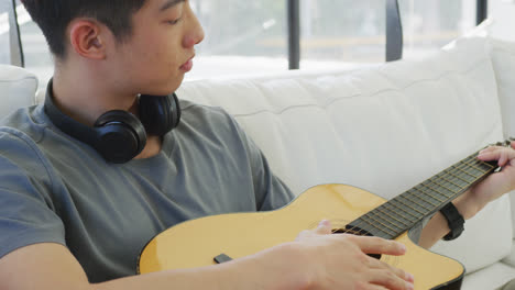 Asiatischer-Männlicher-Teenager-Mit-Kopfhörern,-Der-Gitarre-Spielt-Und-Im-Wohnzimmer-Sitzt