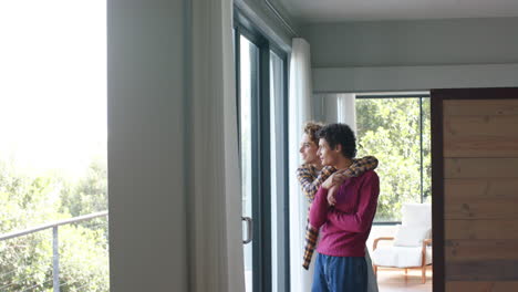 Happy-diverse-gay-male-couple-looking-through-window-and-embracing-at-home,-slow-motion