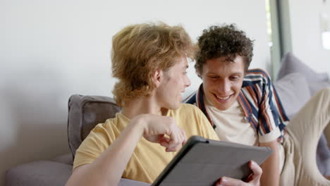 Happy-diverse-gay-male-couple-sitting-at-sofa-using-tablet-at-home,-slow-motion