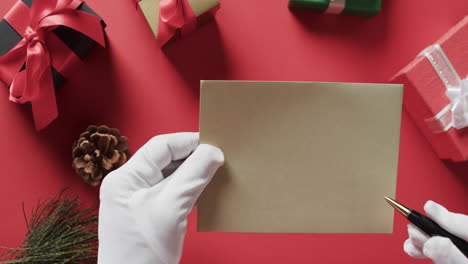 Video-Vom-Weihnachtsmann,-Der-Einen-Braunen-Umschlag-Mit-Kopierraum-Und-Weihnachtsgeschenken-Auf-Rotem-Hintergrund-Hält