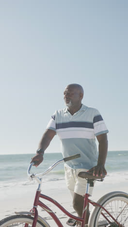 Vertical-video-of-happy-senior-african-american-man-walking-with-bike-at-beach,-in-slow-motion