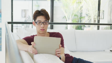 Asiatischer-Männlicher-Teenager-Trägt-Eine-Brille-Und-Benutzt-Ein-Tablet-Im-Wohnzimmer