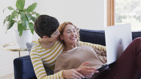 Happy-caucasian-lesbian-couple-lying-on-sofa,-using-laptop-and-laughing-in-sunny-house