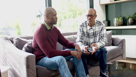 Glücklicher-Afroamerikanischer-Senior-Vater-Und-Sohn-Sitzen-Auf-Der-Couch,-Reden-Und-Lachen,-Zeitlupe