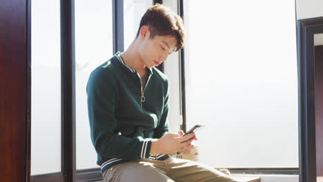 Asiatischer-Junge-Mit-Smartphone-Sitzt-Zu-Hause-Am-Fenster