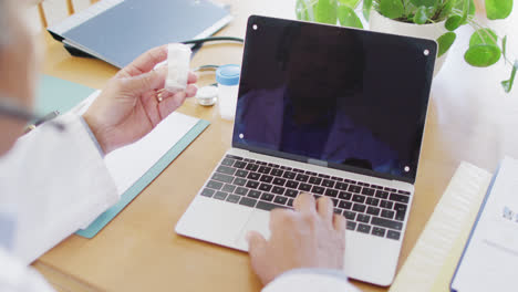 Senior-biracial-male-doctor-using-laptop-and-holding-medication,-copy-space-on-screen