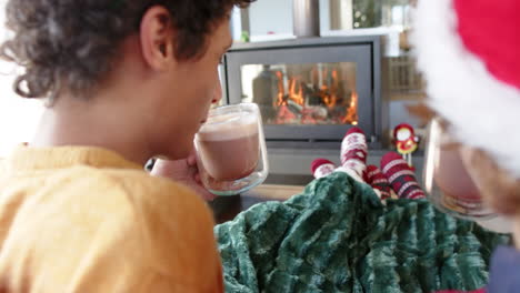 Diverse-gay-male-couple-drinking-cocoa-on-sofa-by-fireplace-at-home-at-christmas,-slow-motion