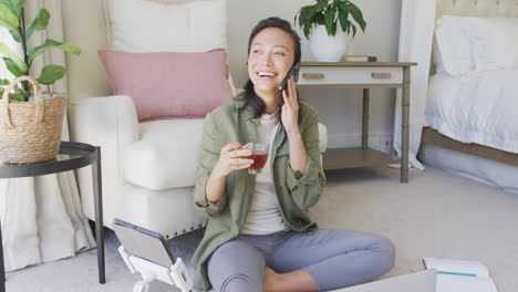 Feliz-Mujer-Asiática-Hablando-En-Un-Teléfono-Inteligente-Y-Bebiendo-Té-En-El-Dormitorio,-En-Cámara-Lenta