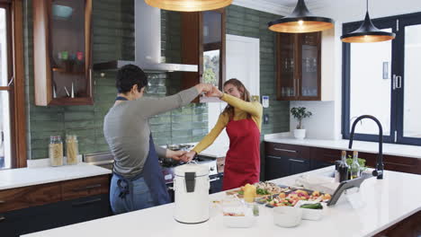 Happy-caucasian-lesbian-couple-preparing-food,-dancing-and-using-tablet-in-sunny-kitchen