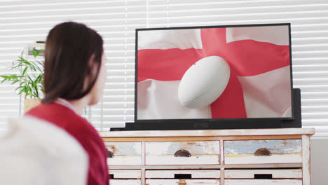 Caucasian-woman-watching-tv-with-rugby-ball-on-flag-of-england-on-screen