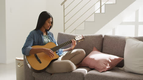 Video-Einer-Glücklichen-Frau-Mit-Gemischter-Abstammung,-Die-Auf-Dem-Sofa-Sitzt-Und-Gitarre-Spielt