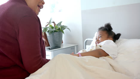 Happy-african-american-mother-fooling-with-daughter-lying-in-hospital-bed-with-mascot,-slow-motion