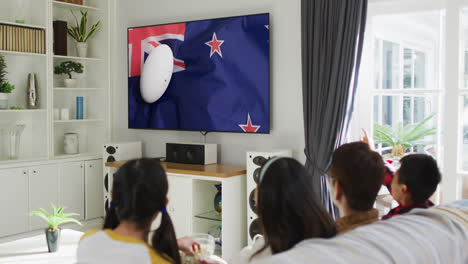 Asian-family-watching-tv-with-rugby-ball-on-flag-of-new-zealand-on-screen