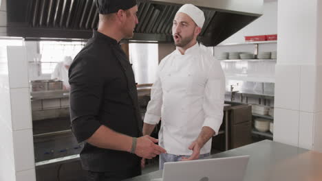 Focused-caucasian-male-chef-instructing-trainee-male-chef-using-laptop-in-kitchen,-slow-motion