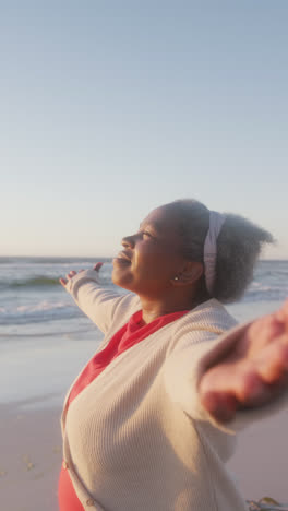 Vertikales-Video-Einer-Glücklichen-älteren-Afroamerikanerin,-Die-Am-Strand-Die-Arme-Ausbreitet,-In-Zeitlupe