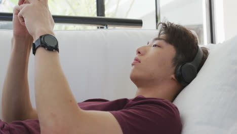 Asiatischer-Männlicher-Teenager-Trägt-Kopfhörer-Und-Benutzt-Smartphone-Im-Wohnzimmer