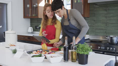 Happy-caucasian-lesbian-couple-preparing-food-and-using-tablet-in-sunny-kitchen