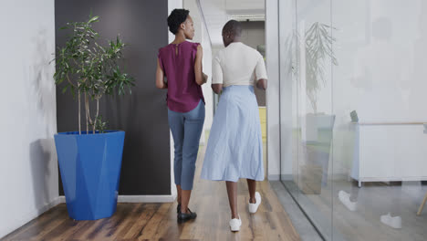 Rear-view-of-two-african-american-businesswomen-talking,-walking-in-office-corridor,-in-slow-motion