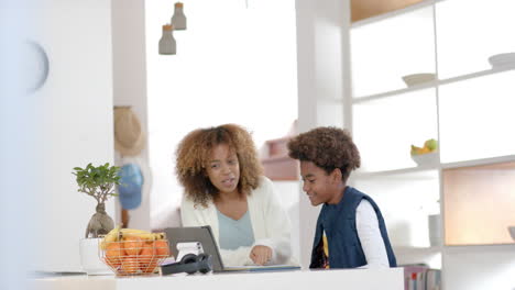 Happy-african-american-mother-helping-her-son-with-homework-using-tablet-in-kitchen,-slow-motion