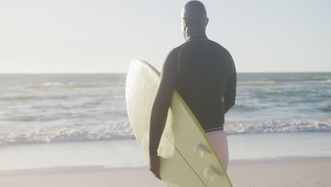 Glücklicher-älterer-Afroamerikanischer-Mann,-Der-In-Zeitlupe-Am-Strand-Spazieren-Geht-Und-Ein-Surfbrett-Hält