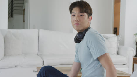 Porträt-Eines-Glücklichen-Asiatischen-Männlichen-Teenagers,-Der-Im-Wohnzimmer-Kopfhörer-Trägt