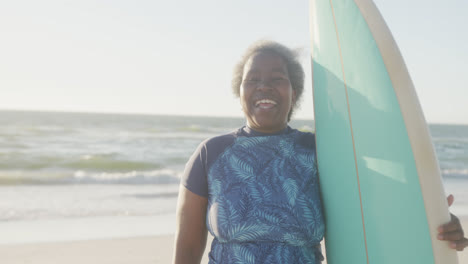 Portrait-of-happy-senior-african-american-woman-holding-surfboard-at-beach,-in-slow-motion