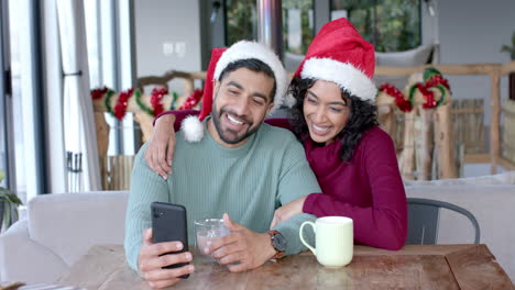 Feliz-Pareja-Birracial-Usando-Sombreros-De-Santa-Claus-Usando-Un-Teléfono-Inteligente-Para-Videollamadas-En-Casa,-Cámara-Lenta