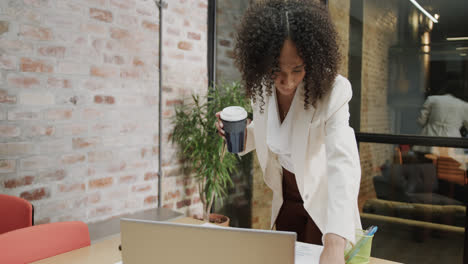 Biracial-businesswoman-with-takeaway-coffee-standing-at-desk-looking-at-document,-in-slow-motion