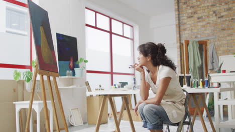 Biracial-woman-ponders-her-painting-in-a-bright-studio