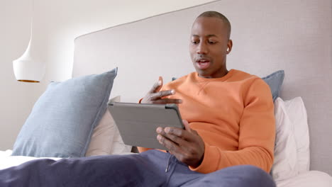 Feliz-Hombre-Afroamericano-Sentado-En-La-Cama-Usando-Una-Tableta-Para-Videollamadas-En-Casa,-Cámara-Lenta