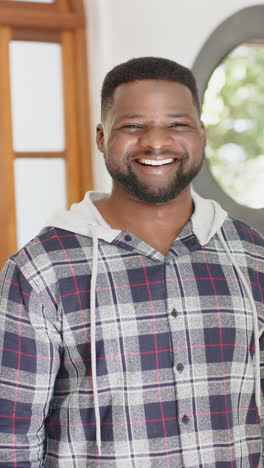 Vertical-video-of-happy-african-american-man-with-beard-standing-and-smiling-in-sunny-house