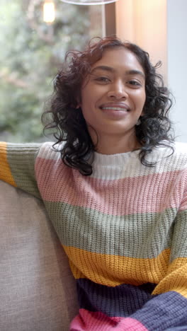 Vertical-video-of-portrait-of-happy-biracial-woman-sitting-on-sofa-at-home,-slow-motion