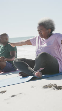 Vertical-video-of-senior-african-american-couple-doing-yoga-at-beach,-in-slow-motion