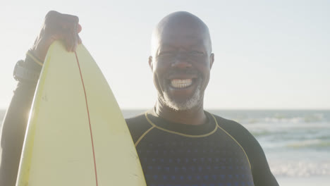 Retrato-De-Un-Feliz-Hombre-Afroamericano-Mayor-Sosteniendo-Una-Tabla-De-Surf-En-La-Playa,-En-Cámara-Lenta