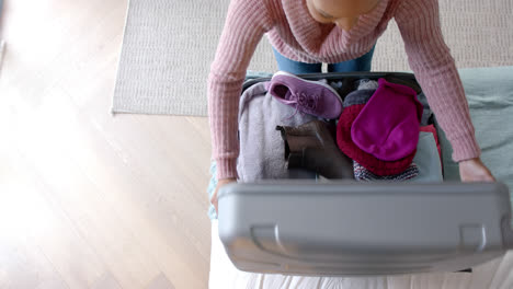 Overhead-view-of-biracial-woman-packing-suitcase-in-sunny-bedroom,-slow-motion